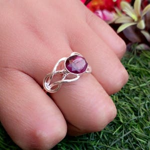 May include: A silver ring with a deep purple oval gemstone set in a delicate, interwoven band.