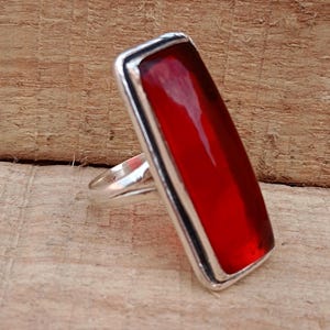 Puede incluir: Un anillo de plata con una piedra roja rectangular. El anillo tiene un diseño sencillo y está engastado en una banda de plata.