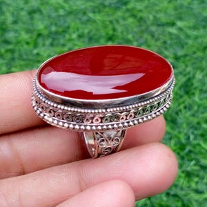 May include: A silver ring with an oval, deep red gemstone. The gemstone is set in a detailed, ornate silver band. The ring's design features intricate patterns and a polished finish, showcasing a classic jewelry style.