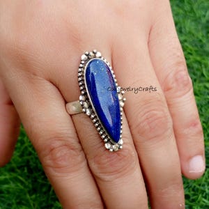 May include: A silver ring featuring a large, teardrop-shaped, deep blue gemstone. The gemstone is surrounded by a detailed, beaded silver setting. The ring is displayed on a hand against a blurred green background. The text "on jewelry crafts" is visible.