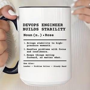May include: A white ceramic mug with a black rim and handle, held by a person. The mug features the text "DEVOPS ENGINEER BUILDS STABILITY" and a definition. The mug's design includes the name "Rose" and a list of qualities.