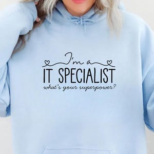 May include: Light blue hoodie with a front pocket and the text "I'm a IT SPECIALIST what's your superpower?" in black. The hoodie has a drawstring hood and long sleeves. The text is in a handwritten style font.