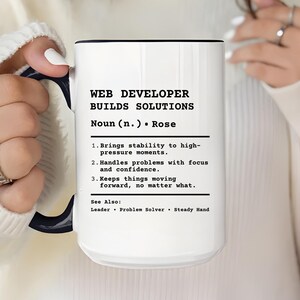 Könnte beinhalten: Weißer Keramikbecher mit schwarzem Innenraum und Henkel, mit dem Text "WEB DEVELOPER BUILDS SOLUTIONS". Der Becher enthält eine Definition eines Webentwicklers, die seine Fähigkeiten und Qualitäten hervorhebt.
