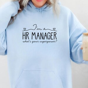 Peut inclure: Sweat à capuche bleu clair avec une poche avant et l'inscription noire "I'm a HR MANAGER what's your superpower ?". Le sweat à capuche a une capuche à cordon et des manches longues.
