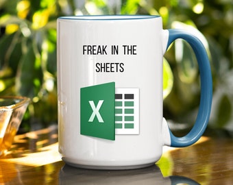 May include: White ceramic mug with a blue handle and rim. The mug features the text "FREAK IN THE SHEETS" above an Excel logo. A clear glass bowl with liquid is in the foreground.