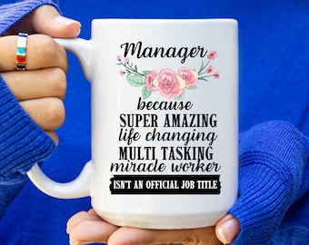 May include: White ceramic mug with the text "Manager" above a floral design and the phrases "SUPER AMAZING", "life changing", "MULTI TASKING", and "miracle worker". The text "ISN'T AN OFFICIAL JOB TITLE" is also included.