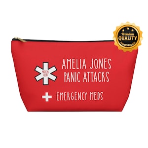 Puede incluir: Un estuche rojo con cremallera con el texto "AMELIA JONES PANIC ATTACKS EMERGENCY MEDS". Incluye un símbolo de alerta médica blanco y un sello dorado de "Premium Quality".