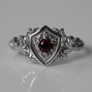 May include: A silver ring featuring a central shield-shaped setting with an intricate filigree design on the band. A single round, deep red gemstone is set in the middle of the shield, which is surrounded by detailed metal scrollwork patterns.