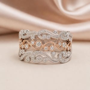 May include: Three ornate rings with a mix of silver and rose gold tones. The rings feature a swirling, filigree design, accented with numerous small, sparkling diamonds. The rings are stacked, showcasing their intricate details and elegant style.