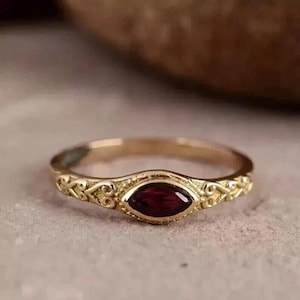 May include: A gold-colored ring with a maroon-colored, oval-shaped stone at the center. The ring has an ornate, filigree design on the band. The ring is set on a neutral-colored surface.