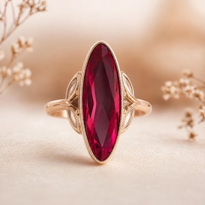 May include: A gold-toned ring featuring a large, elongated, faceted ruby gemstone. The ruby is a deep red color and is set in a gold band with decorative filigree details on either side of the gemstone. The ring is displayed on a neutral background.