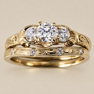 May include: A gold engagement ring set with a three-stone design. The center stone is a large, round diamond, flanked by two smaller diamonds. The band features intricate floral engravings and additional small diamonds. The set includes a matching wedding band.