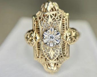 May include: A gold filigree ring with a central, flower-shaped setting. The ring features intricate openwork and two decorative side accents. The center setting holds a small, clear gemstone. The ring is displayed on a white surface.