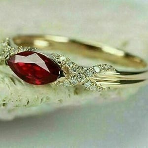 May include: A gold-toned ring featuring a marquise-cut, deep red gemstone as the focal point. The band is adorned with small, clear accent stones. The ring's design incorporates a delicate, intertwined pattern, creating an elegant and sophisticated look.