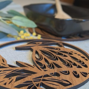 Könnte beinhalten: Holzuntersetzer mit detailliertem, geschichtetem Design. Der Untersetzer hat eine dunkelbraune Kontur und komplizierte Ausschnittmuster, die die natürliche Holzmaserung zeigen. Das Design scheint ein stilisierter Vogel oder ein Blumenmotiv zu sein.