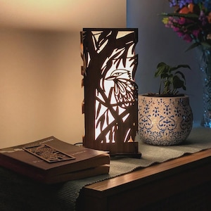 May include: A wooden table lamp with a bird design cut into the front. The lamp is rectangular and has a warm glow. A book and a decorative bookmark are on the table. A potted plant and flowers are also on the table.