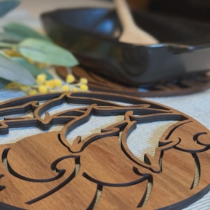 Könnte beinhalten: Holz-Topfuntersetzer mit einem lasergeschnittenen Design von schwimmenden Delfinen. Der Untersetzer ist aus hellbraunem Holz mit einem dunkelbraunen Rand gefertigt. Eine schwarze Schüssel und ein Holzlöffel sind im Hintergrund, mit Grün.