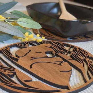 Może przedstawiać: Drewniana podstawka z motywem misia koala. Podstawka wykonana jest z ciemnobrązowego drewna i posiada szczegółowe wycięcie misia koala. Czarna ceramiczna miska i drewniana łyżka znajdują się w tle. Średnica podstawki wynosi około 10 cm.