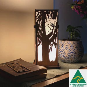 Puede incluir: Lámpara de madera con diseño de koala, con una silueta de árbol. La lámpara es rectangular y se mantiene en posición vertical. Un libro y un marcapáginas decorativo están sobre la mesa. Una planta en maceta y flores en el fondo.