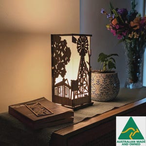 May include: A wooden lamp featuring a farmhouse and windmill silhouette, emitting a warm light. The lamp is rectangular and placed on a wooden surface, alongside a book with a decorative bookmark. A vase of flowers and a potted plant are in the background.