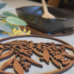 Könnte beinhalten: Ein hölzerner Untersetzer mit detailliertem Blumendesign. Der Untersetzer ist kreisförmig mit einer dunkelbraunen Oberfläche. Das Design zeigt Blätter und kleine Blütentrauben. Eine dunkle Schüssel und ein Holzlöffel befinden sich im Hintergrund.