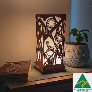 May include: A wooden lamp with a nature-themed design. The lamp features a bird and floral cutouts, with a warm light emanating from within. The lamp is rectangular and sits on a wooden base. A book and a potted plant are nearby.