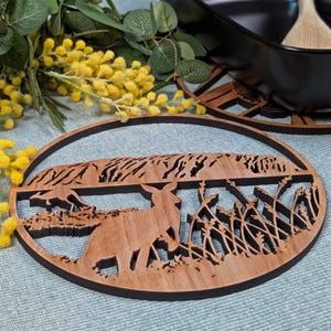 Könnte beinhalten: Ovaler Holzuntersetzer mit einem lasergeschnittenen Design, das einen Hirsch in einem Feld zeigt. Der Untersetzer hat einen dunkelbraunen Rand und eine hellbraune Innenseite. Das Design umfasst hohes Gras und eine entfernte Landschaft.