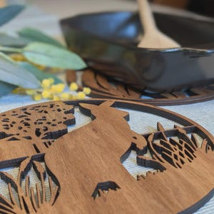 Może przedstawiać: Drewniana podstawka z sylwetką kangura, drzewami i wyciętym wzorem trawy. Podstawka wykonana jest z brązowego drewna i ma okrągły kształt. Czarna patelnia żeliwna i drewniana łyżka znajdują się w tle.