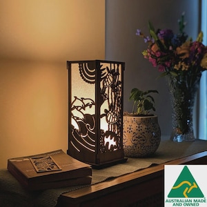 May include: A wooden lamp with a rectangular frame and detailed cut-out patterns of waves, trees, and turtles. The lamp emits a warm light, highlighting the intricate designs. A book and bookmark are placed nearby.