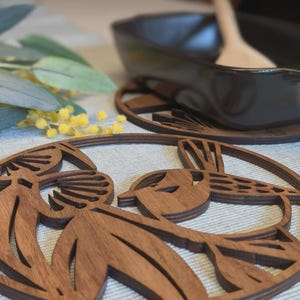 May include: Wooden coasters with a bird design. The coasters are made of dark brown wood and feature a laser-cut bird motif. A black dish and wooden spoon are in the background, along with yellow and green floral accents. The coasters are circular.