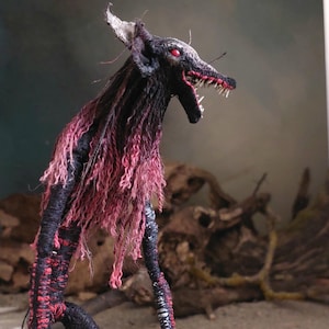 May include: A handcrafted sculpture of a creature with a wolf-like head and elongated limbs. The sculpture features a black and red color scheme, with red eyes and a mane of pinkish-red fibers. The creature has an open mouth revealing sharp teeth.