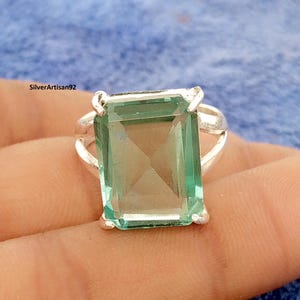 Può includere: Un anello d'argento con una grande pietra preziosa rettangolare verde chiaro. La pietra preziosa è incastonata in una fascia d'argento con un design a gambo diviso. L'anello è mostrato su uno sfondo blu.