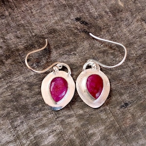 Könnte beinhalten: Ein Paar silberne Ohrringe mit einem tropfenförmigen, tiefroten Edelstein, der in eine silberne Lünette gefasst ist. Die Ohrringe haben ein einfaches Haken-Design für durchbohrte Ohren. Der Hintergrund ist eine strukturierte Holzoberfläche.