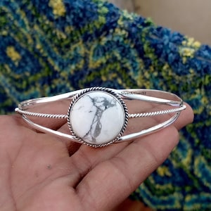 Könnte beinhalten: Ein silbernes Armreif-Armband mit einem großen, runden weißen Stein mit grauer Maserung. Der Stein ist in einer gedrehten, seilartigen Bordüre gefasst. Das Armband hat ein schlichtes, elegantes Design. Der Text "SilverArtisan92" ist sichtbar.