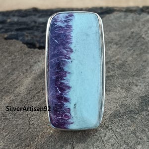 Può includere: Un anello d'argento con una pietra rettangolare di colore viola e blu. La pietra preziosa presenta un motivo unico di viola e blu.