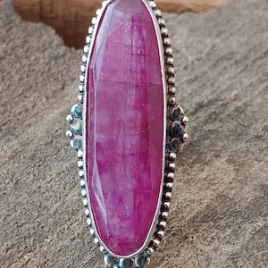 May include: A sterling silver ring featuring a large, oval-cut ruby gemstone. The ruby is a vibrant magenta color, set in a bezel with a beaded border. The ring has decorative silver accents on the sides.