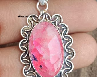 Op de afbeelding: Ovale roze edelsteen hanger met een zilveren filigraan frame. De hanger heeft een beugel voor een ketting. De edelsteen heeft een mix van roze en paarse tinten. De hanger wordt door een hand vastgehouden.