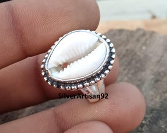 May include: A silver ring with a white cowrie shell set in the center. The ring is decorated with a beaded border.
