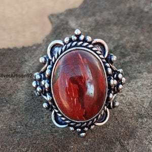 May include: A silver ring with a red and white agate stone set in an ornate oval bezel. The ring has a detailed design with small beads and a rope-like border.