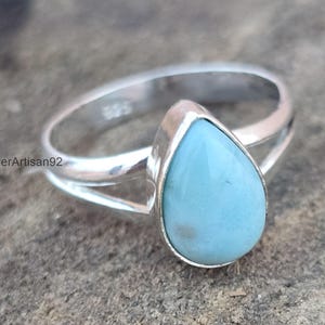 Könnte beinhalten: Ein silberner Ring mit einem tropfenförmigen blauen Edelstein. Der Ring ist in einer einfachen Band mit einer geteilten Schiene gefasst.