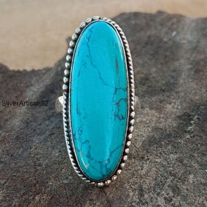May include: A turquoise oval ring with a silver band and beaded edging. The ring is set on a rough, brown surface.