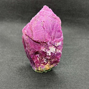 May include: A raw ruby gemstone in a deep magenta hue. The surface has a textured, crystalline appearance with lighter pink and white patches. The base has a touch of green. The gemstone is set against a dark grey background.