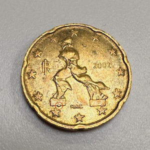 May include: A gold-coloured coin with a detailed design. The coin features a depiction of a person with a flowing cape, surrounded by stars. The year "2002" is visible. The coin has the letters "R" and "MAC" on it.