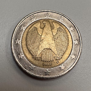 Op de afbeelding: Een 2 euro munt uit 2002 met een gedetailleerd adelaarsontwerp in het midden, omringd door een goudkleurige binnenring en een zilverkleurige buitenring. De munt heeft een sterrenrand.