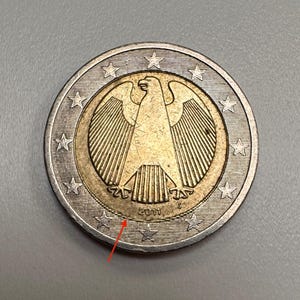 May include: A 2011 two-euro coin with the German eagle design. The coin has a gold centre and a silver outer ring. Twelve stars are spaced around the edge.