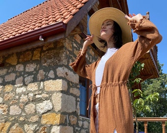 May include: A brown linen long-sleeved dress with a tie waist and a front slit. The dress is worn with a white vest top and a straw hat.