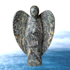Peut inclure: Une figurine d'ange en pierre avec des ailes déployées. La statue est principalement grise avec des accents bruns et verts, et a une surface texturée. L'ange se tient debout, les mains jointes devant sa poitrine. L'arrière-plan est une image floue de l'océan.