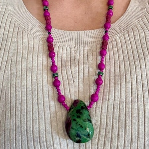 Op de afbeelding: Een opvallende ketting met een grote, groen en rode stenen hanger. De ketting heeft een kralenontwerp met fuchsia-kleurige kralen en accenten. De ketting wordt gedragen over een neutraal gekleurde top.