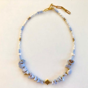 May include: A delicate necklace featuring a mix of white and light blue beads, accented with light blue gemstone chips and a gold charm.