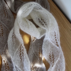 Pode incluir: Close-up de duas echarpes de malha, uma bege clara e outra creme. Ambas apresentam um design vazado com um padrão de folhas. As echarpes estão drapeadas sobre uma superfície de madeira. Acessório de moda.